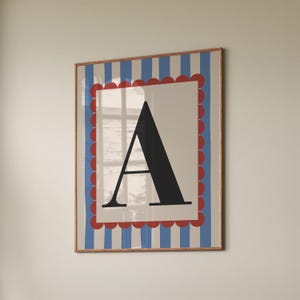 Puede incluir: Un estampado artístico enmarcado con una gran letra "A" negra sobre un fondo crema. La obra de arte tiene un borde festoneado rojo y rayas verticales azules y blancas. El marco es de color marrón claro.