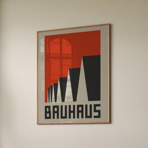 Könnte beinhalten: Ein gerahmter Druck mit rotem Hintergrund und einem geometrischen Design in Schwarzweiß. Das Design zeigt eine Reihe von Dreiecken, die eine Gebäudeform bilden. Der Text "Bauhaus" ist unten auf dem Druck in Weiß gedruckt.