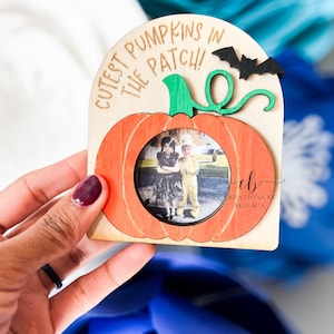 Puede incluir: Un marco de fotos de madera con forma de calabaza con un recorte para una foto. El marco está pintado de naranja y tiene un tallo verde y un murciélago negro. El texto "Cutest Pumpkins in the Patch!" está grabado en el marco.