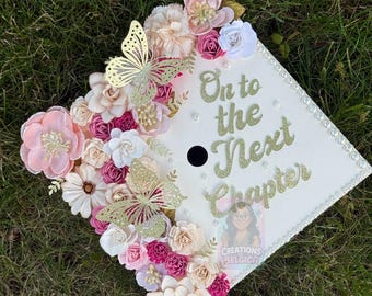May include: A decorated graduation cap with the words "On to the Next Chapter" in gold glitter. The cap is adorned with an assortment of fabric flowers in shades of pink, white, and cream, along with gold butterfly accents.