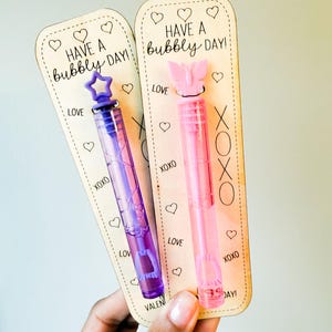 May include: Two bubble wands with wooden backing cards. One wand is purple with a star-shaped top, the other is pink with a butterfly top. The cards read "Have a bubbly day!" with hearts and "Love" and "XOXO" text.