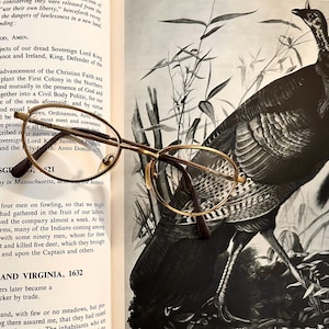 May include: Gold-rimmed eyeglasses with oval lenses rest on an open book. The book's pages display historical text. A detailed illustration of a turkey stands in the background, adding a vintage aesthetic.