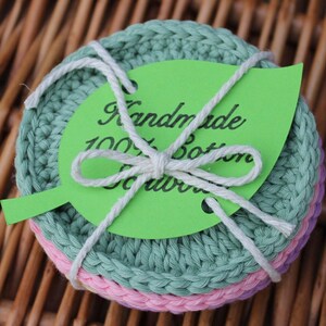 May include: A stack of handmade cotton scrubbers in shades of green, pink, and purple, tied with white string. A green leaf-shaped tag reads "Handmade 100% Cotton Scrubbers" in black script. The scrubbers are crocheted.