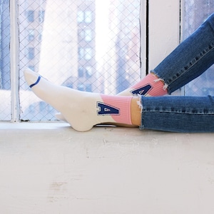 May include: A pair of white socks with pink and blue accents. The socks feature a blue "A" on a pink background. The socks are worn by a person wearing blue jeans.