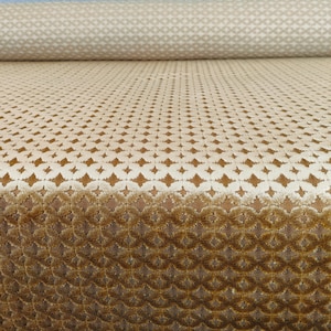 Può includere: Un rotolo di tessuto beige con un motivo a rombi ripetuto. Il tessuto ha un aspetto strutturato, simile al velluto, con un gradiente di colore da chiaro a scuro. Il motivo è un mix di forme geometriche.