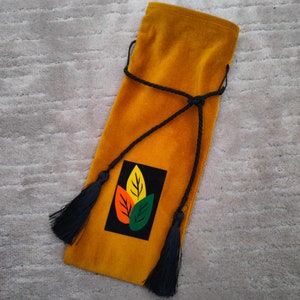 Puede incluir: Una bolsa de vino de terciopelo amarillo mostaza con un cord&oacute;n y un borla negros. La bolsa tiene un parche negro y blanco con un dise&ntilde;o de hojas de colores.