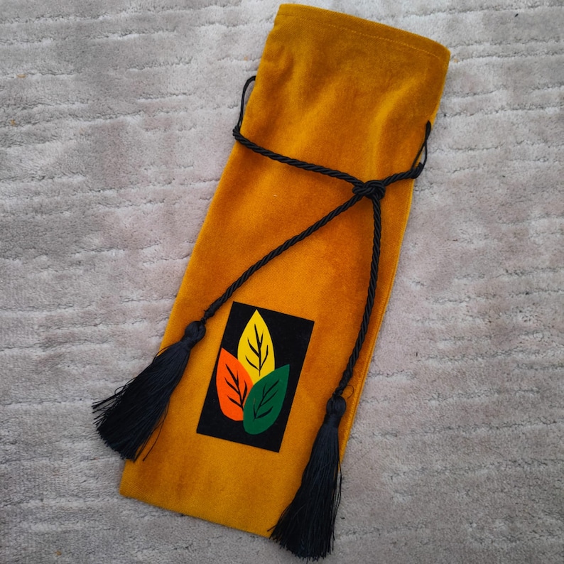 Puede incluir: Una bolsa de vino de terciopelo amarillo mostaza con un cord&oacute;n y un borla negros. La bolsa tiene un parche negro y blanco con un dise&ntilde;o de hojas de colores.