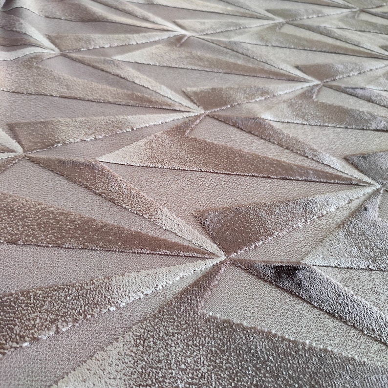 Op de afbeelding: Close-up van een beige stof met een geometrisch patroon van reli&euml;f driehoeken. De stof heeft een zacht, getextureerd oppervlak.