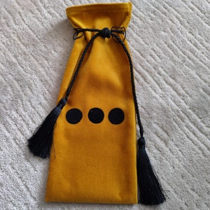 Puede incluir: Una bolsa de cord&oacute;n de terciopelo amarillo mostaza con borlas negras y tres c&iacute;rculos negros en la parte delantera.