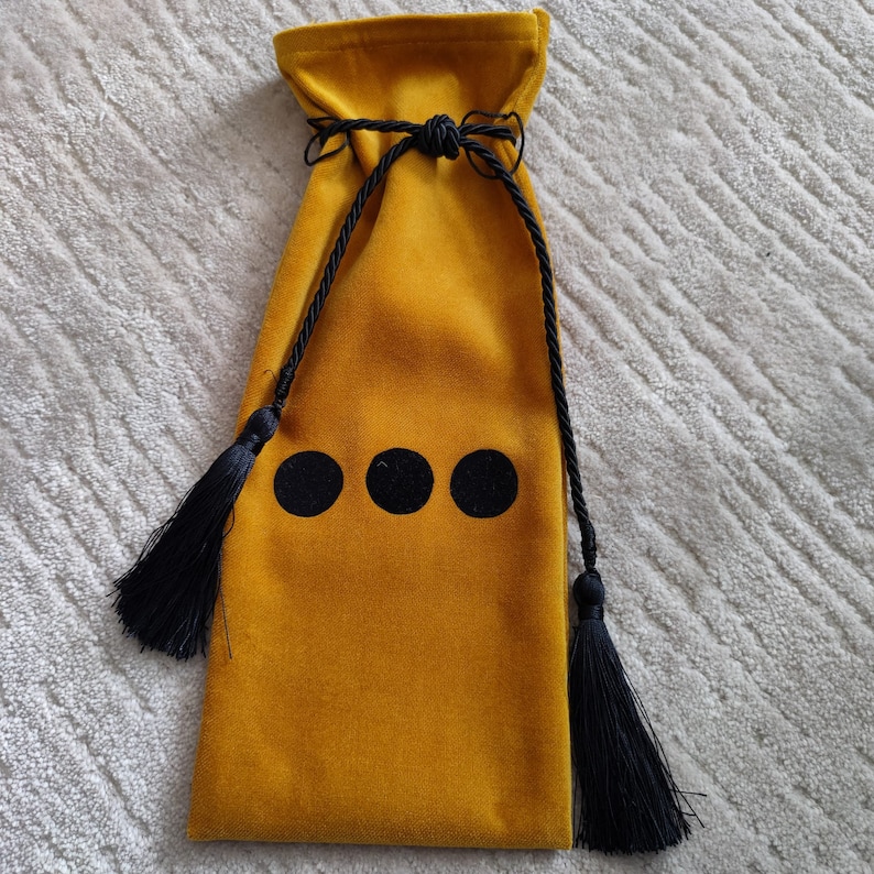 Puede incluir: Una bolsa de cord&oacute;n de terciopelo amarillo mostaza con borlas negras y tres c&iacute;rculos negros en la parte delantera.