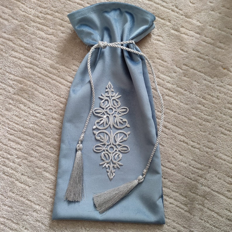Puede incluir: Una bolsa de cord&oacute;n de terciopelo azul claro con un dise&ntilde;o bordado blanco. La bolsa tiene dos borlas plateadas.