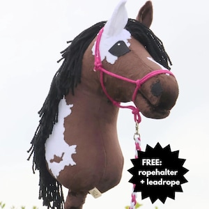 May include: A brown and white hobby horse with black mane and a pink rope halter. The toy horse has a wooden stick handle and a white and pink lead rope. The image includes the text "FREE: ropehalter + leadrope".