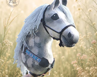 May include: A grey dapple hobby horse with a long light grey yarn mane, dark grey fabric ears, and black eyes. It features a black rope bridle and a decorative neck strap with a blue, white, and black geometric pattern. Text reads SMarti Horses.