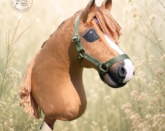 Può includere: Un cavallo a bastone marrone con una macchia bianca sul muso, occhi scuri e criniera in lana marrone chiaro. È dotato di una cavezza in nylon verde oliva con fibbie in metallo ed è fissato a un bastone di legno. Logo circolare e testo visibili.