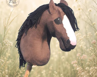 May include: A brown hobby horse with a black yarn mane, a white blaze on its face, and a black muzzle. The horse head is attached to a wooden stick. A circular logo in the top left corner reads SMarti Horses, and text at the bottom reads Handmade in Czech Republic.