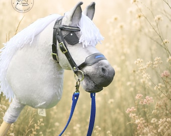 May include: A light gray plush hobby horse with a white yarn mane, featuring a black leather bridle with gold-tone buckles, a blue noseband detail, and a blue fabric rein. The horse has a circular logo on the neck and a small tag on the side.