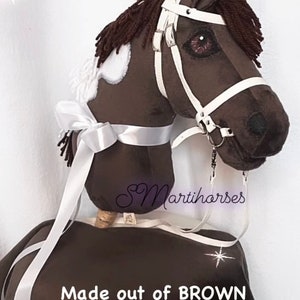 Op de afbeelding: Een bruin en wit hobbypaard met een wit hoofdstel en een wit lint. Het paard is gemaakt van bruin velboa-stof en heeft een witte ster aan de zijkant. De tekst "Made out of BROWN Velboa shade" staat onder het paard.