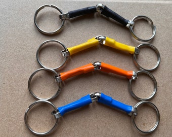 Puede incluir: Cuatro bocados de caballo de diferentes colores con anillos plateados. Los bocados son negro, amarillo, naranja y azul.
