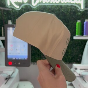 May include: A beige baseball cap with a green brim. The cap has the word "OUT" embroidered on the side in green thread. The cap is being held by a hand with red nail polish.