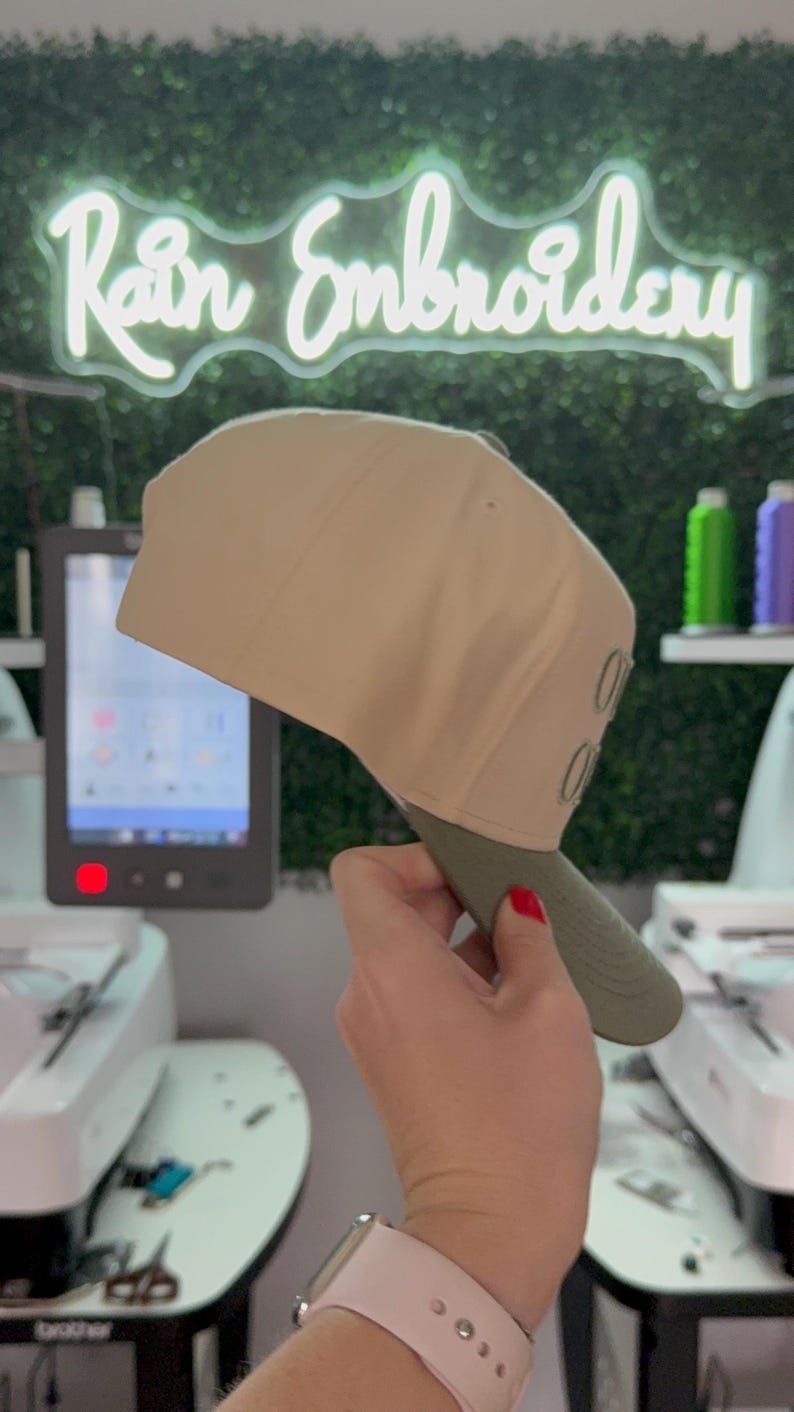 May include: A beige baseball cap with a green brim. The cap has the word "OUT" embroidered on the side in green thread. The cap is being held by a hand with red nail polish.