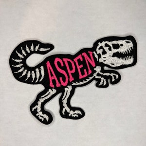 May include: A black and white embroidered patch of a dinosaur skeleton with the word "ASPEN" written in pink across the body.