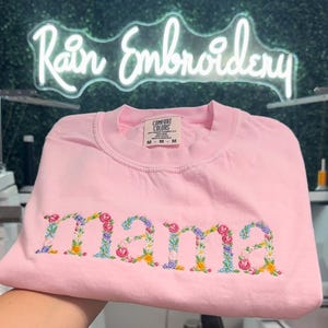 K&ouml;nnte beinhalten: Rosafarbenes T-Shirt mit dem Wort "mama" in bunten Blumen gestickt. Das Shirt ist gefaltet und hat ein Etikett mit der Aufschrift "Comfort Colors" und "M - M - M".