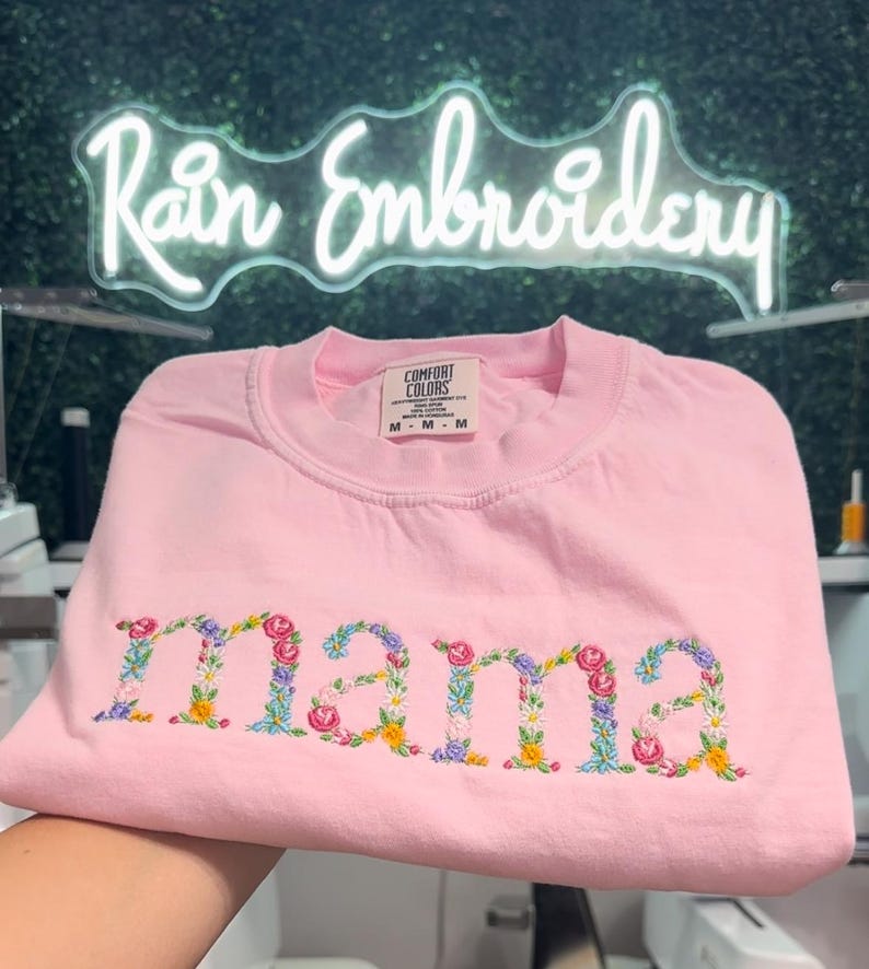 K&ouml;nnte beinhalten: Rosafarbenes T-Shirt mit dem Wort "mama" in bunten Blumen gestickt. Das Shirt ist gefaltet und hat ein Etikett mit der Aufschrift "Comfort Colors" und "M - M - M".