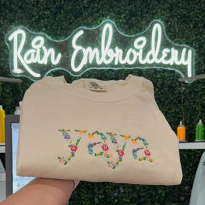 K&ouml;nnte beinhalten: Ein cremefarbenes T-Shirt mit dem Wort "YOURS" in bunten Blumen gestickt.
