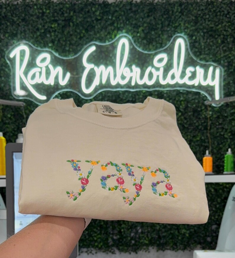 K&ouml;nnte beinhalten: Ein cremefarbenes T-Shirt mit dem Wort "YOURS" in bunten Blumen gestickt.