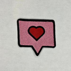 Może przedstawiać: Haftowany naszywka w kolorze różowym i czarnym z czerwonym sercem w środku dymku.