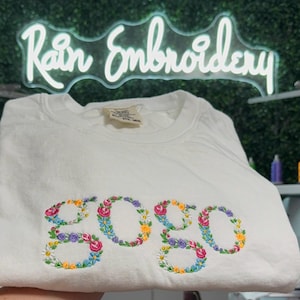 K&ouml;nnte beinhalten: Wei&szlig;es T-Shirt mit dem Wort "gogo", das mit bunten Blumen bestickt ist.