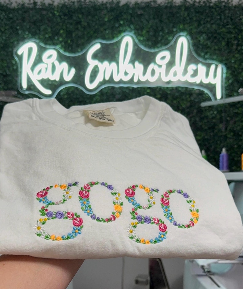 K&ouml;nnte beinhalten: Wei&szlig;es T-Shirt mit dem Wort "gogo", das mit bunten Blumen bestickt ist.