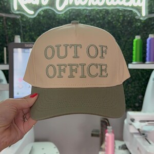 May include: A beige and green baseball cap with the text "OUT OF OFFICE" embroidered in white on the front.