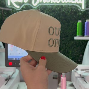 May include: A beige and green baseball cap with the text "OUT OF OFFICE" embroidered on the front.