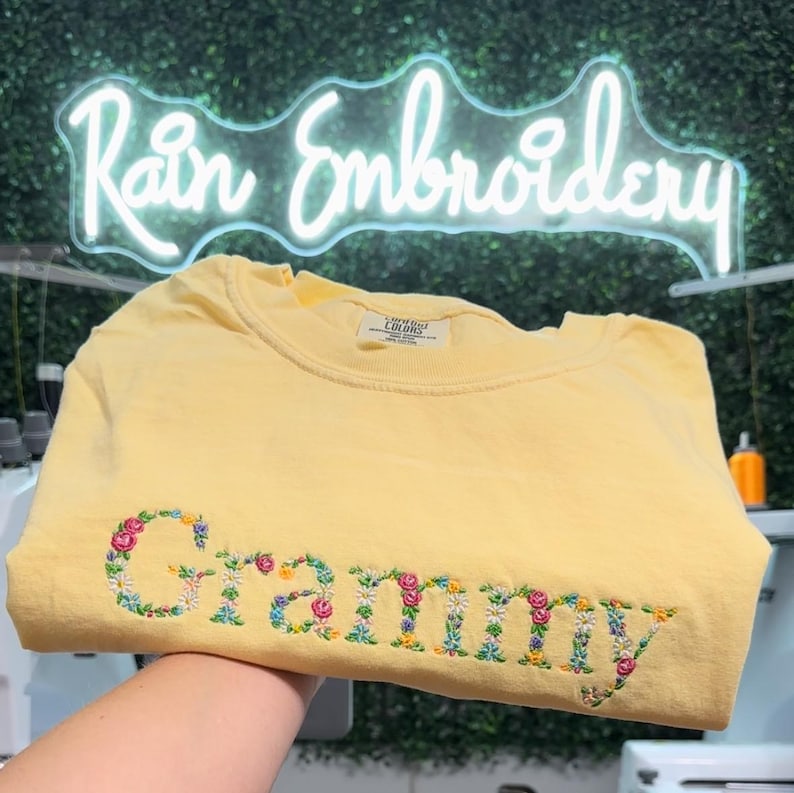 K&ouml;nnte beinhalten: Ein gelbes T-Shirt mit dem Wort "Grammy", das mit bunten Blumen bestickt ist.