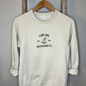 Può includere: Una felpa beige chiaro con un design ricamato nero che dice "Cape Cod est. 1602 Massachusetts" con un'ancora e dettagli floreali.