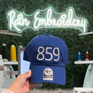 Può includere: Un cappello da baseball blu con il numero 859 ricamato in bianco sul davanti. Il cappello ha un logo 47 bianco sul retro con il testo "47 Clean Up Adjustable".