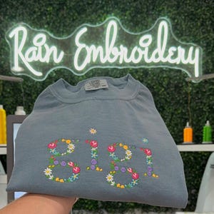 K&ouml;nnte beinhalten: Ein hellblaues T-Shirt mit dem Namen "Gigi", der mit bunten Blumen bestickt ist. Das Shirt ist gefaltet und wird in einer Hand gehalten. Der Hintergrund ist eine gr&uuml;ne Wand mit einem Neon-Schild, auf dem "Rain Embroidery" steht.