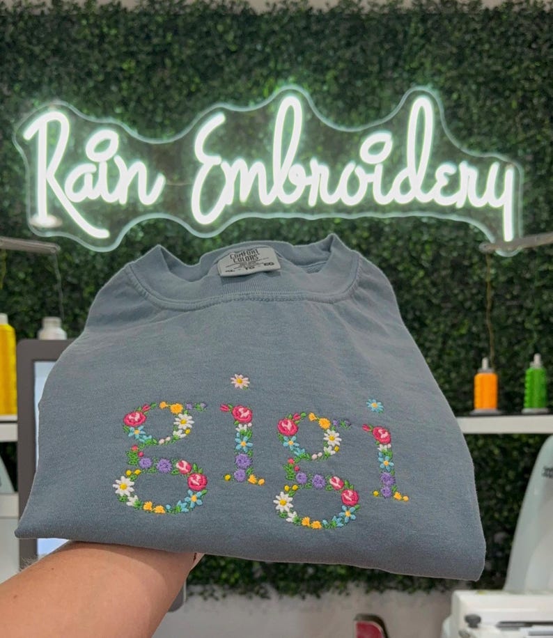 K&ouml;nnte beinhalten: Ein hellblaues T-Shirt mit dem Namen "Gigi", der mit bunten Blumen bestickt ist. Das Shirt ist gefaltet und wird in einer Hand gehalten. Der Hintergrund ist eine gr&uuml;ne Wand mit einem Neon-Schild, auf dem "Rain Embroidery" steht.