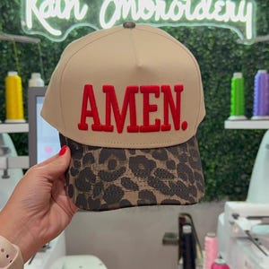 May include: A tan baseball cap featuring a leopard print brim and the word "AMEN" embroidered in red. The cap is held up in front of a neon sign that says "Rain Embroidery".
