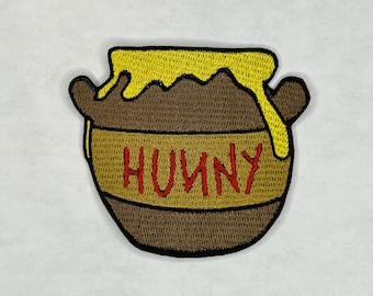 May include: A brown honey pot with a yellow lid and dripping honey. The pot has the word "HUNNY" written on it in red.