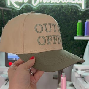 May include: A beige and green baseball cap with the text "OUT OF OFFICE" embroidered in green on the front.