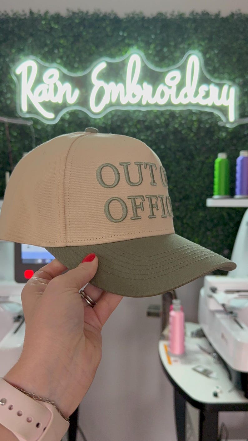 May include: A beige and green baseball cap with the text "OUT OF OFFICE" embroidered in green on the front.