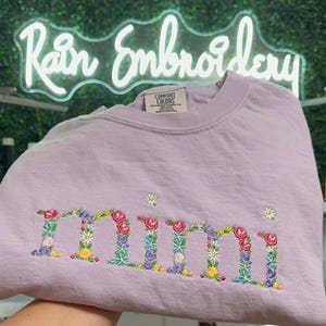 K&ouml;nnte beinhalten: Ein lavendelfarbenes T-Shirt mit dem Wort "mimi", das mit bunten Blumen bestickt ist.