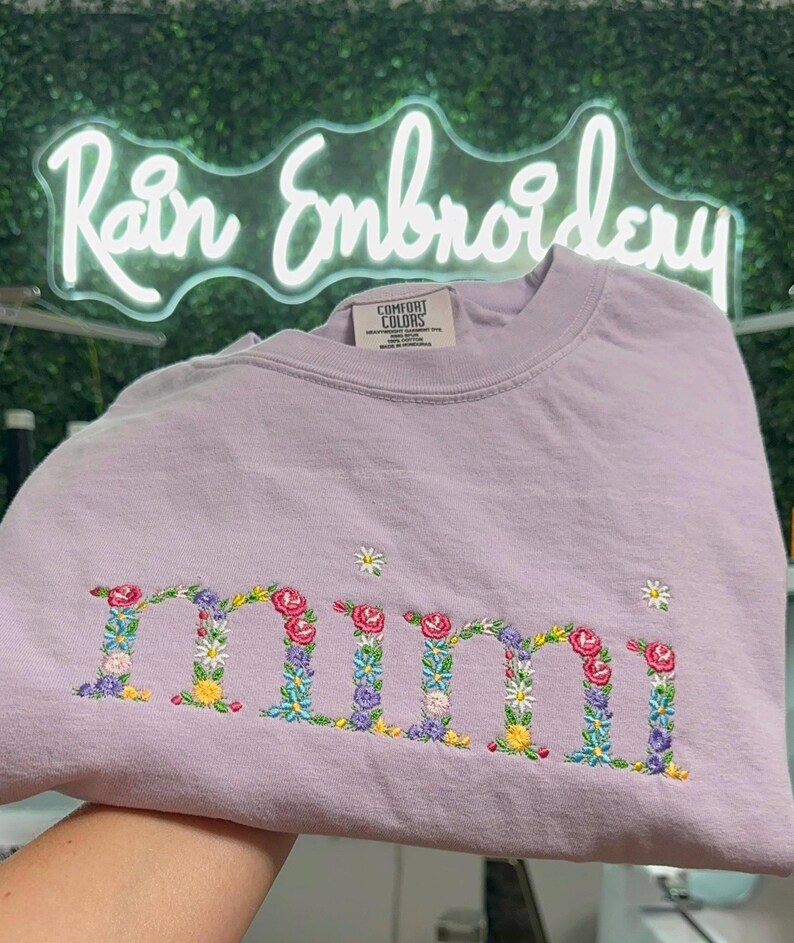 K&ouml;nnte beinhalten: Ein lavendelfarbenes T-Shirt mit dem Wort "mimi", das mit bunten Blumen bestickt ist.