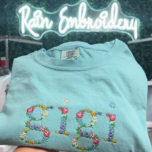 K&ouml;nnte beinhalten: Ein hellblaues T-Shirt mit dem Namen "Gigi", der mit bunten Blumen bestickt ist. Das Shirt ist in der H&auml;lfte gefaltet und wird von einer Hand gehalten. Der Hintergrund ist eine gr&uuml;ne Wand mit einem Neon-Schild, auf dem "Rain Embroidery" steht.