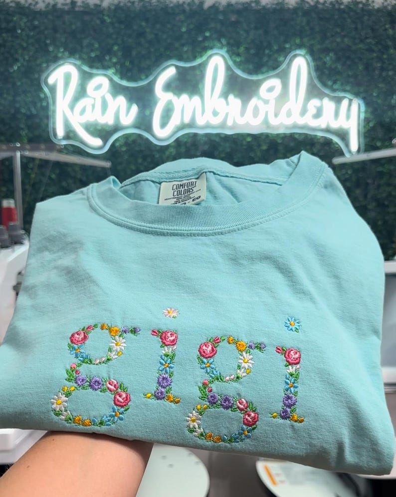 K&ouml;nnte beinhalten: Ein hellblaues T-Shirt mit dem Namen "Gigi", der mit bunten Blumen bestickt ist. Das Shirt ist in der H&auml;lfte gefaltet und wird von einer Hand gehalten. Der Hintergrund ist eine gr&uuml;ne Wand mit einem Neon-Schild, auf dem "Rain Embroidery" steht.