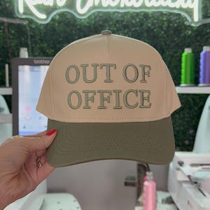 May include: A beige and green baseball cap with the text "OUT OF OFFICE" embroidered in green on the front.