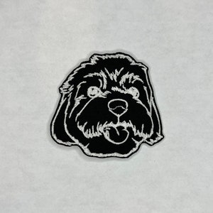 May include: Black and white embroidered patch of a dog's face. The dog has a fluffy, curly coat and is looking directly at the viewer with its tongue sticking out.