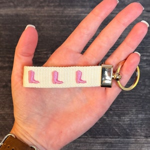 May include: A white key chain with a gold ring and three pink boot charms. The key chain is made of woven fabric.